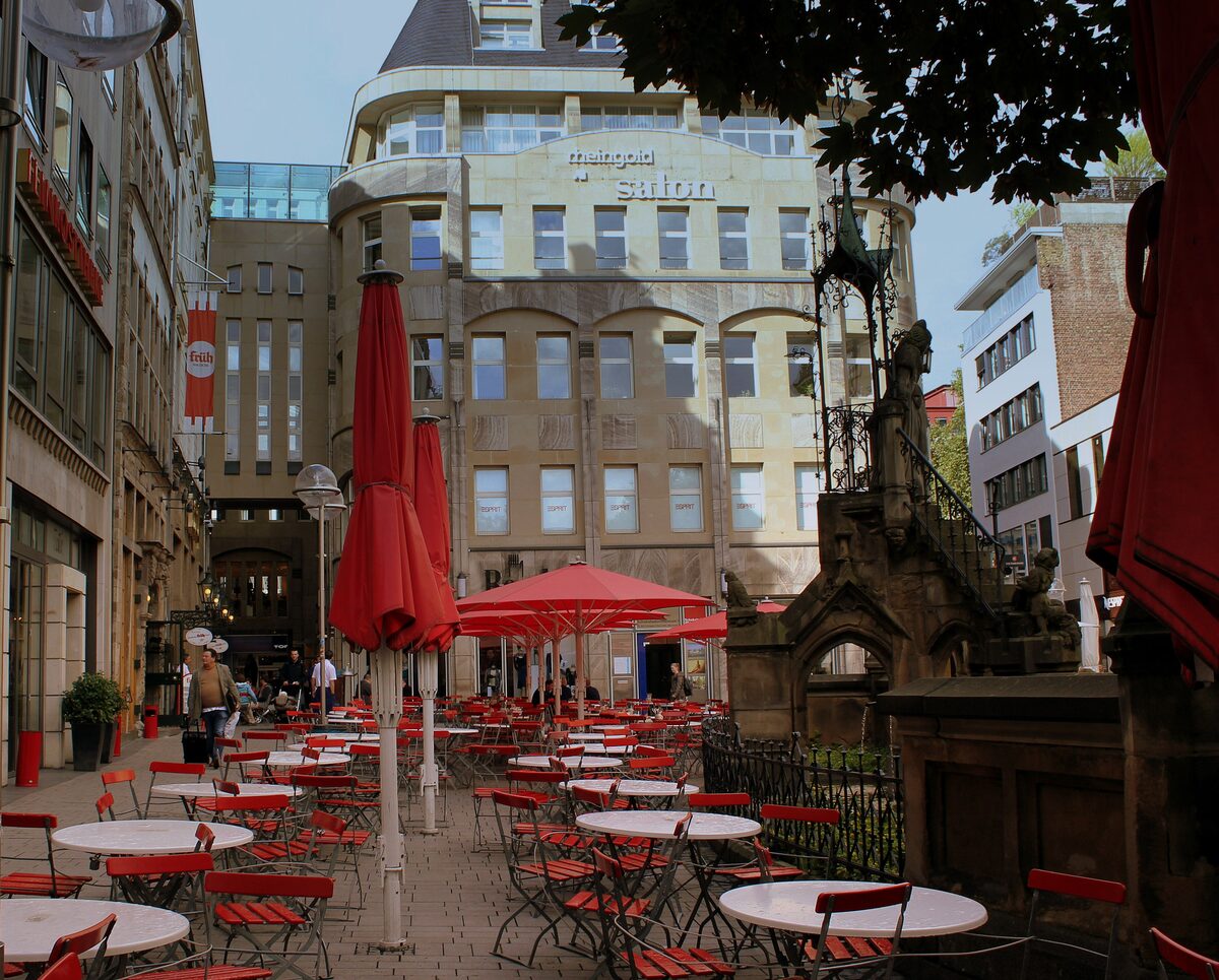 Red parasols and café tables on the terrace of Früh am Dom, with the Heinzelmännchen fountain at the right