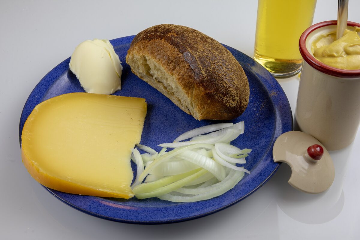 A Halve Hahn deconstructed on a plate: a rye roll, butter, a wedge of Gouda, sliced onion and a pot of mustard, with a glass of Kölsch behind