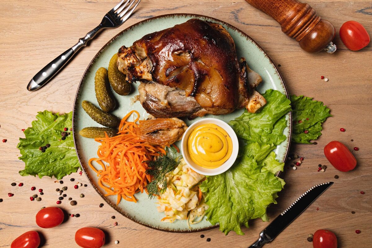 A roast pork knuckle on a plate with pickles, mustard, shredded carrot and lettuce, seen from above