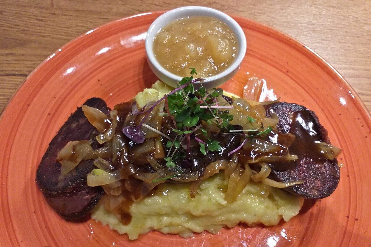 Fried blood sausage on mashed potatoes with fried onions, served with a bowl of apple sauce on an orange plate