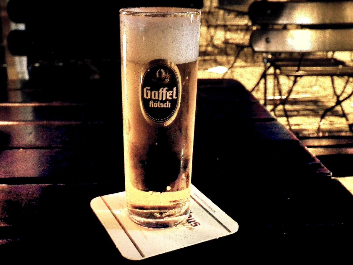 A tall straight glass of Kölsch with a Gaffel logo on a beer mat at a beer garden table