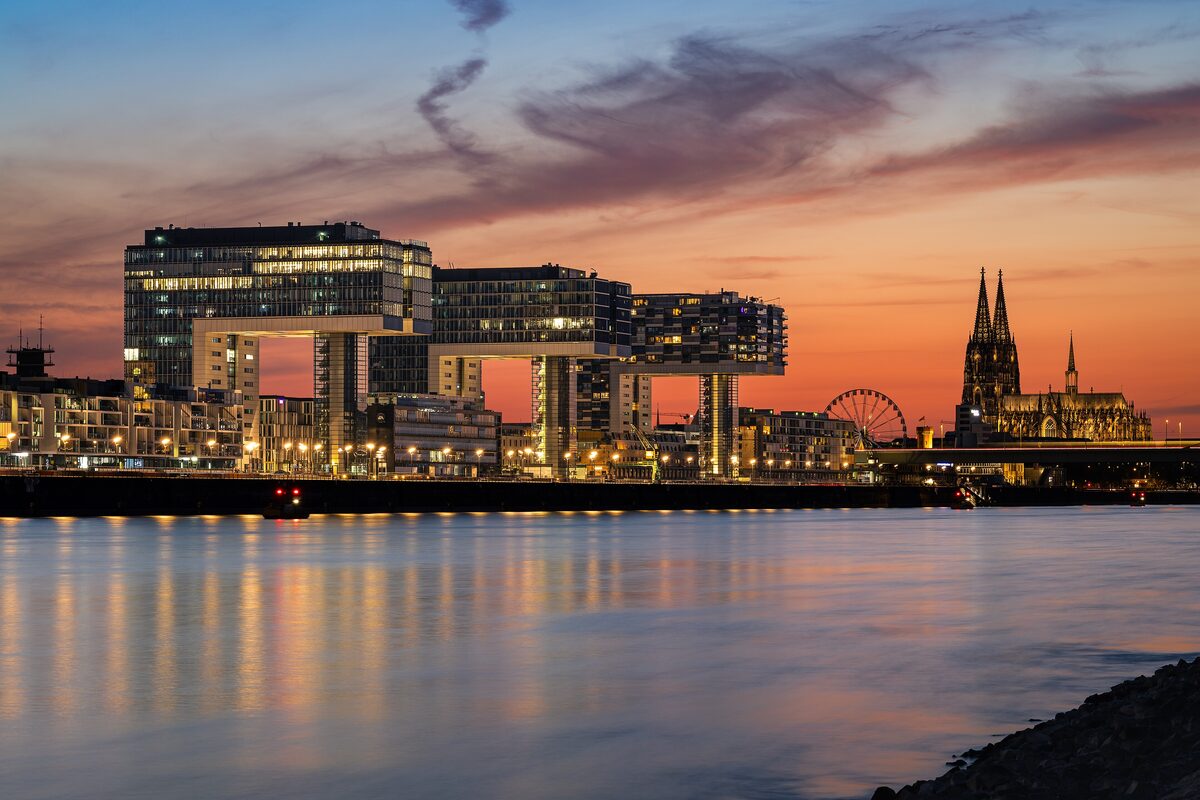 The three Crane Houses lit up at sunset on the Rhine, with the cathedral far right
