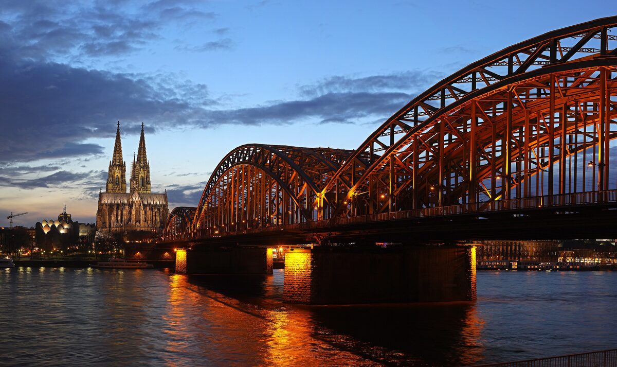 The lit steel arches of the Hohenzollern Bridge and the twin spires of the cathedral at dusk, reflected in the Rhine