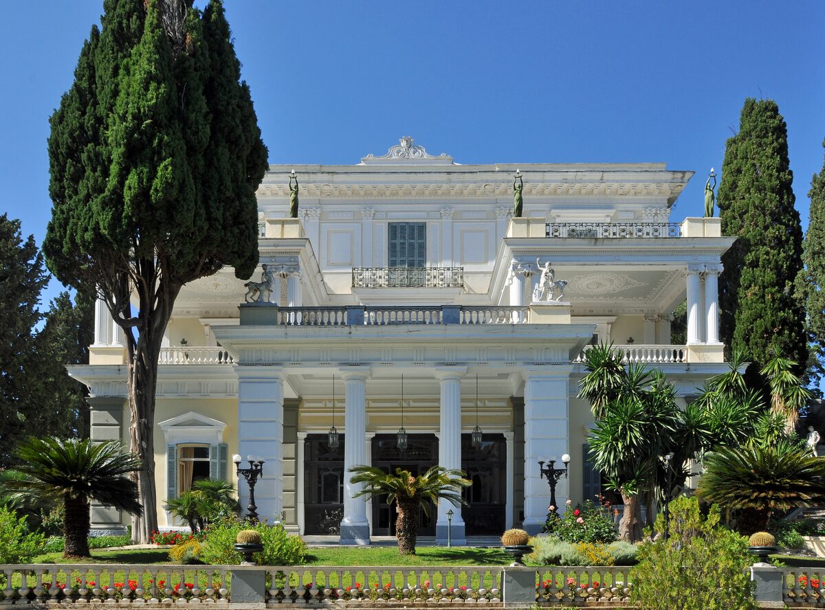The white neoclassical Achilleion palace with its columned porch, behind tall cypresses, palms and a garden railing