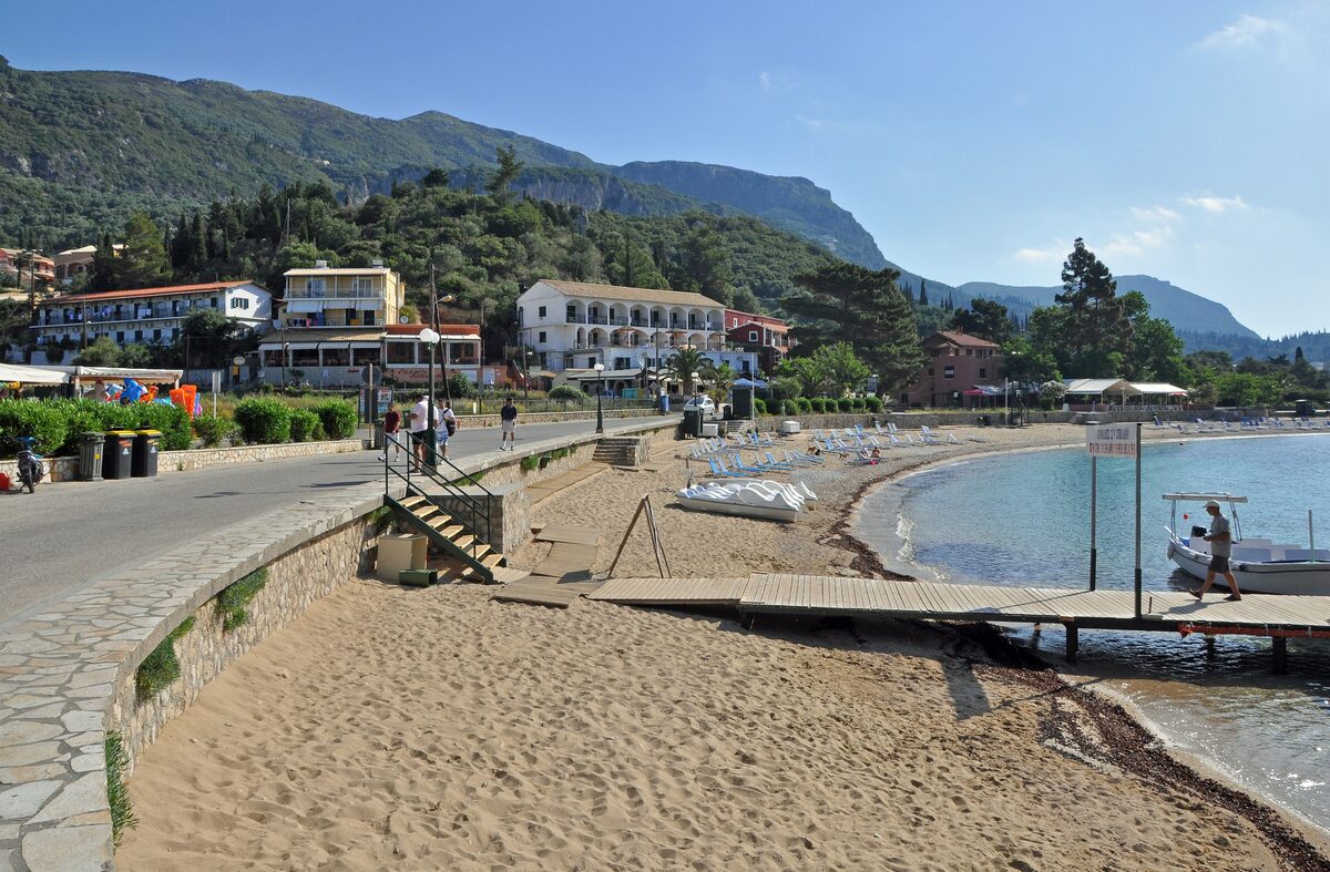 The sandy main beach of Paleokastritsa on a sunny morning, with a wooden jetty, a moored boat, the seafront road and hotels below green hills