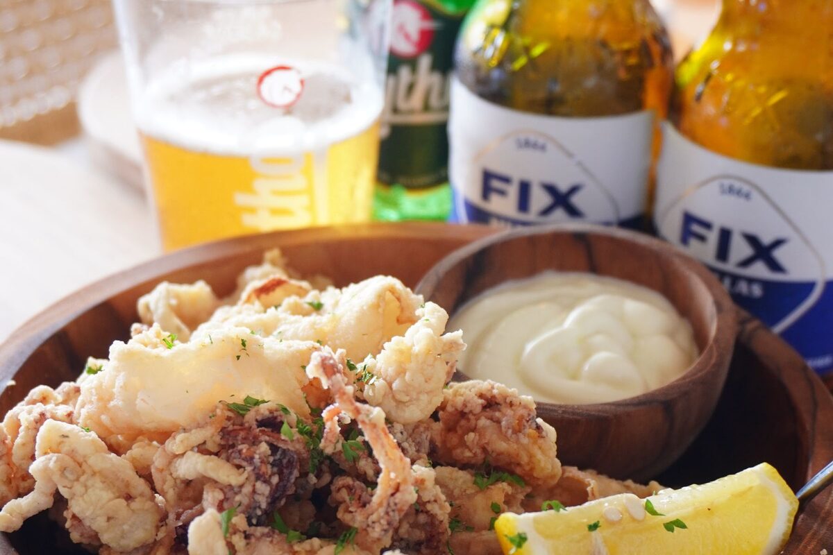 A wooden bowl of fried calamari with lemon and mayonnaise, next to glasses of Greek beer