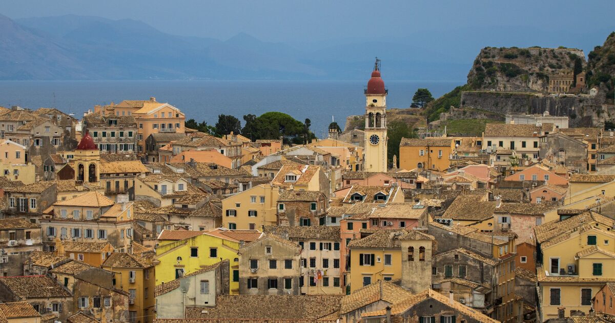 The rooftops of Corfu's old town in warm light, with the red-domed bell tower of St. Spyridon in the middle and the Old Fortress on its rock behind, against the mountains of the mainland