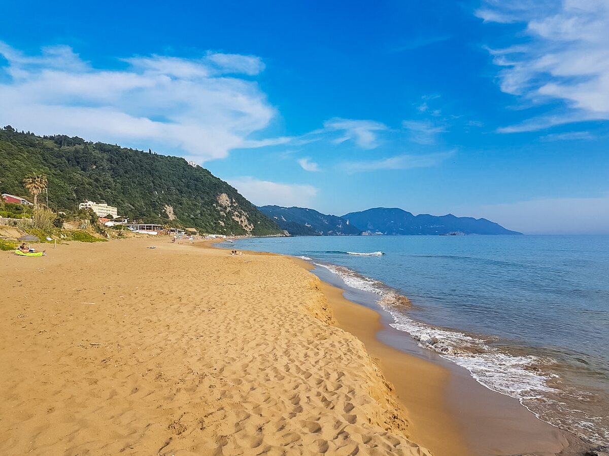 A long stretch of golden sand at Glyfada with waves breaking on the right, wooded hills at the far end and a deep blue sky with white clouds