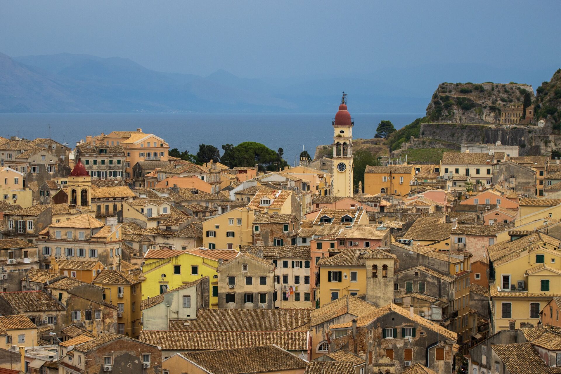 The rooftops of Corfu's old town in warm light, with the red-domed bell tower of St. Spyridon in the middle and the Old Fortress on its rock behind, against the mountains of the mainland
