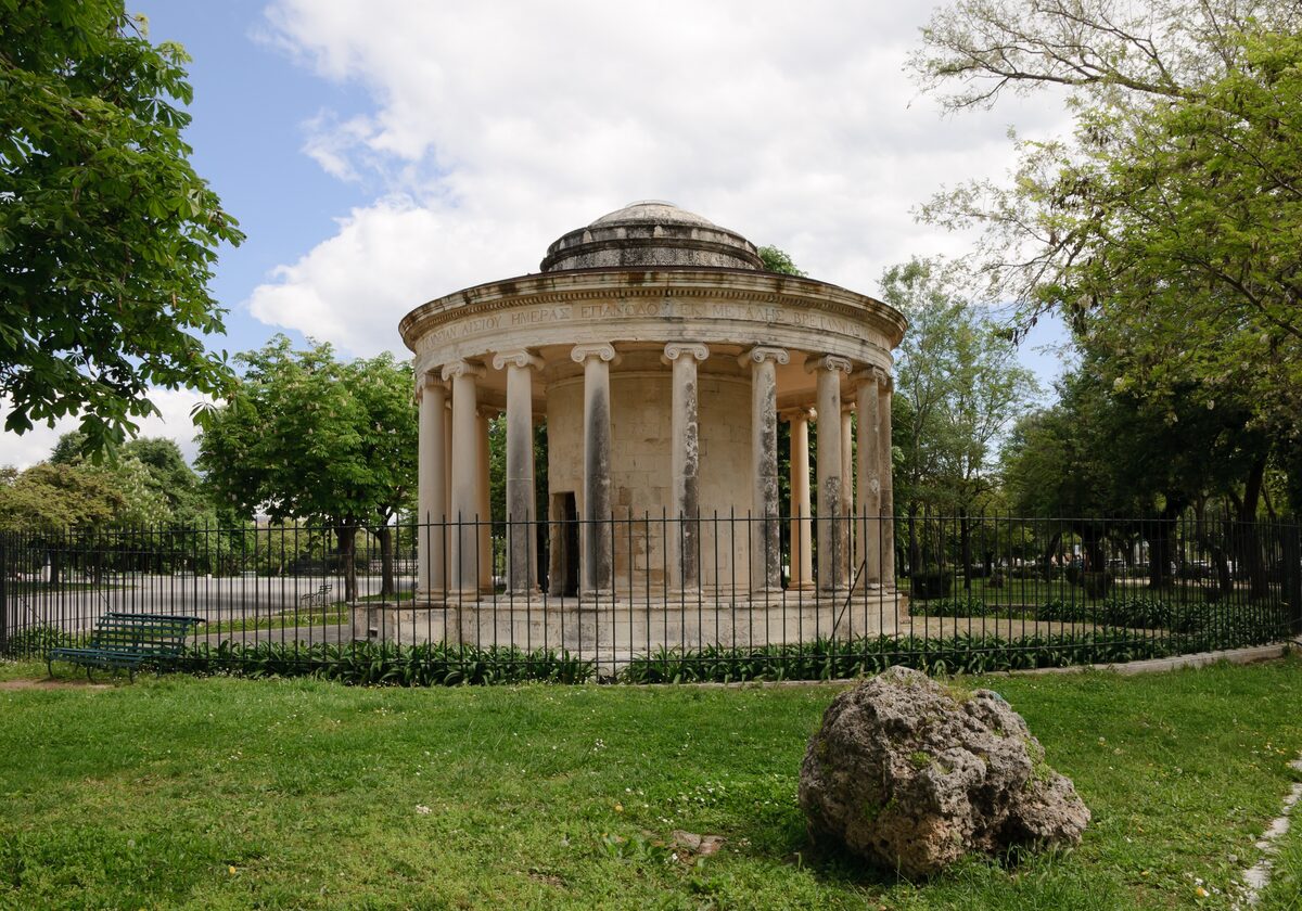 The Maitland Rotunda, a round classical temple of Ionic columns in a fenced garden on the Spianada, under trees