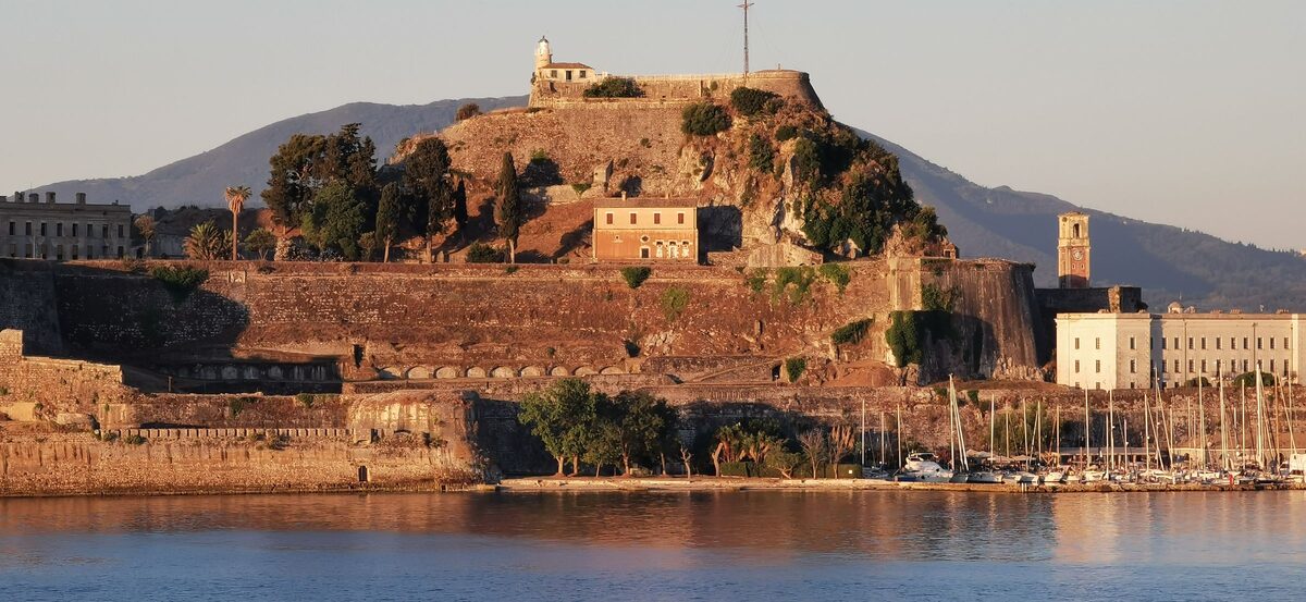 The Old Fortress of Corfu glowing in late afternoon light, its walls and twin peaks rising above the harbor with sailing boats