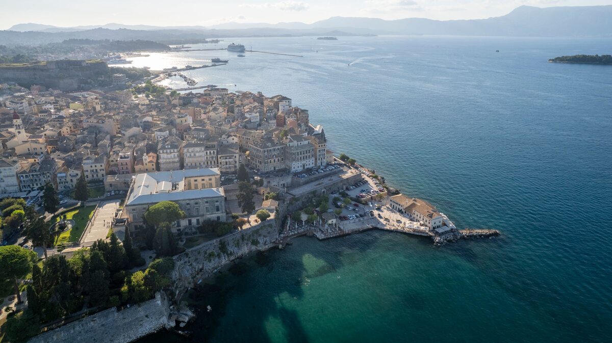 Aerial view of Corfu's old town on its peninsula, a dense block of tall pastel buildings above the sea walls, with the bay and mountains beyond