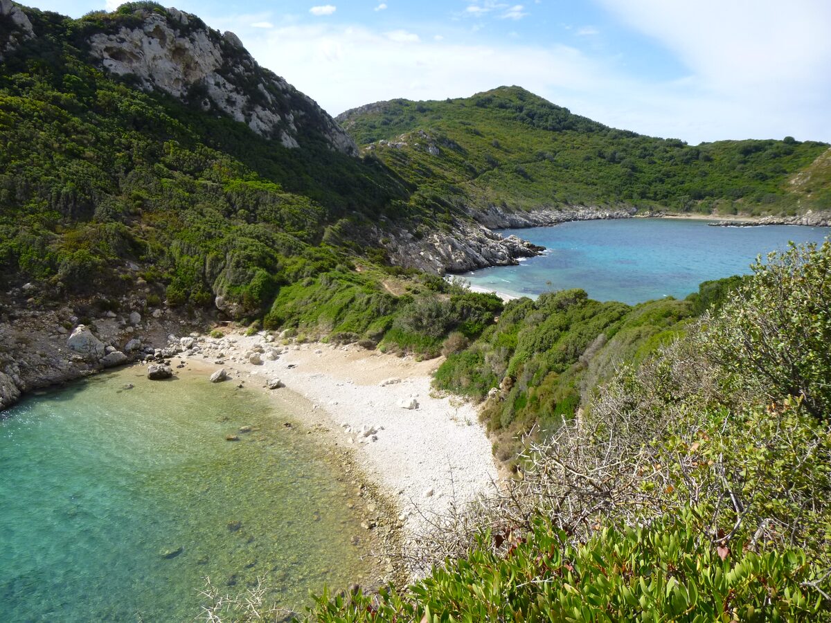 Porto Timoni seen from above: a pale pebble cove with clear turquoise water, and the second bay beyond a green headland under a blue sky
