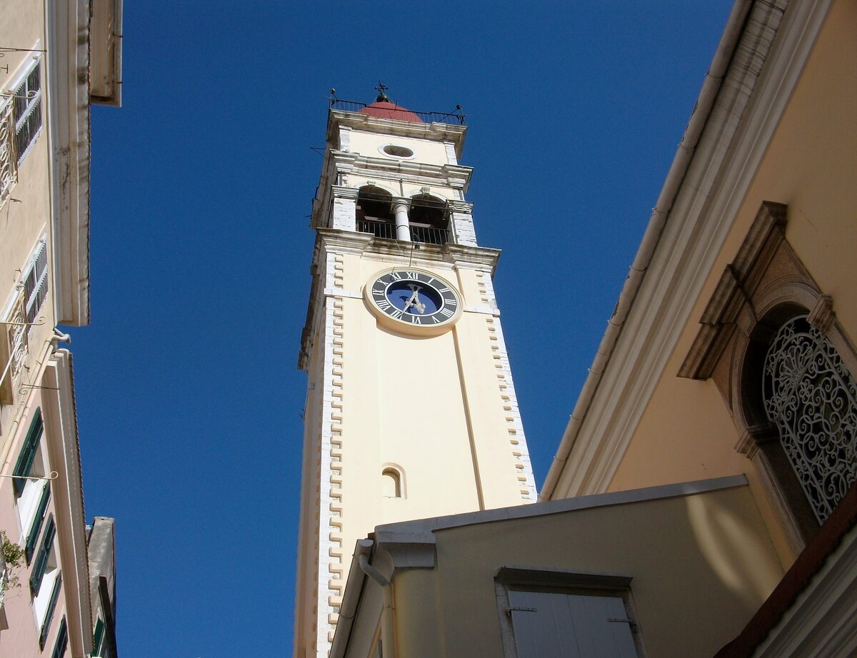 The tall pale bell tower of St. Spyridon church with its clock and red dome, seen from a narrow lane against a deep blue sky