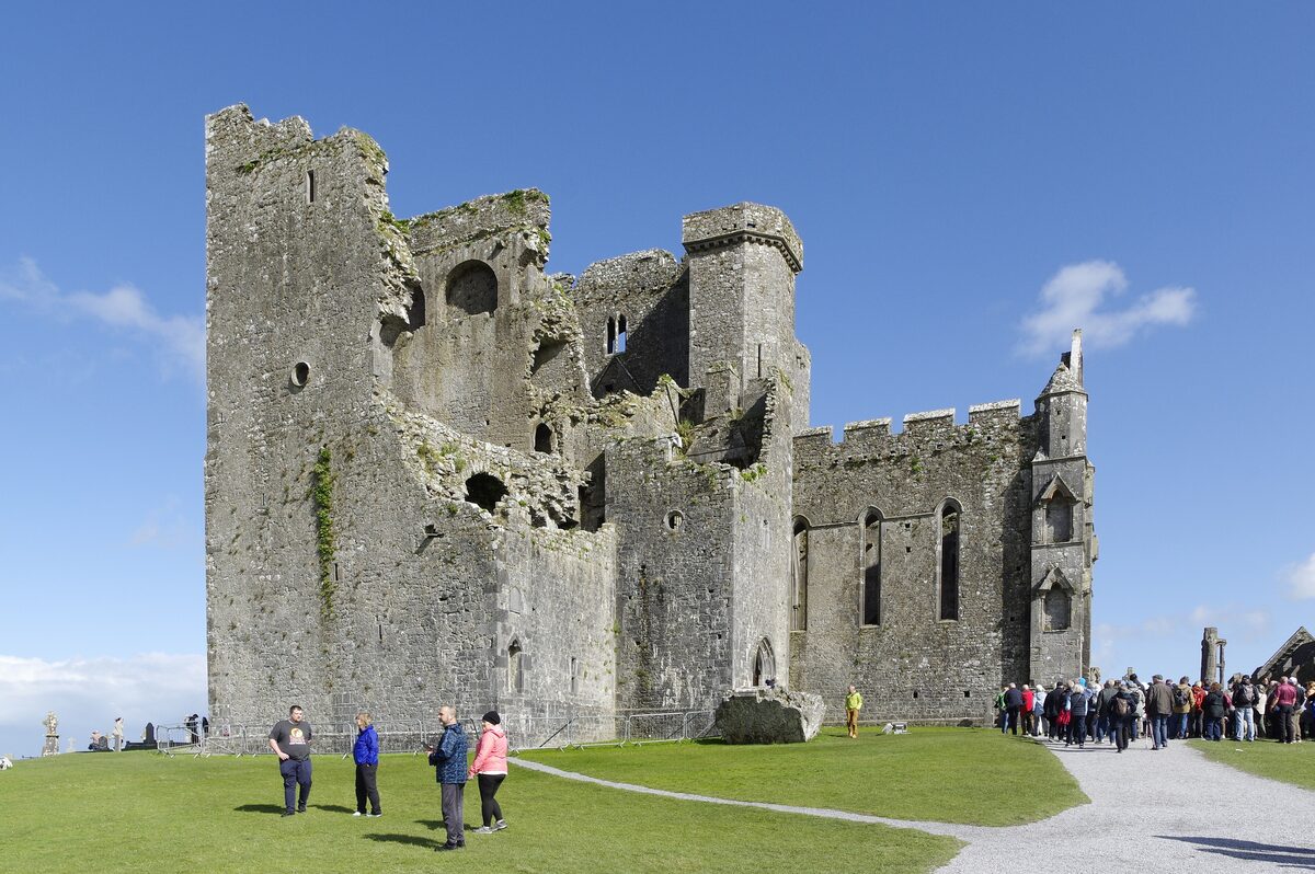 The ruined cathedral and towers of the Rock of Cashel on a green hilltop under a blue sky, with visitors on the path