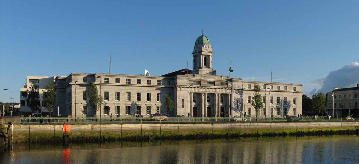 Cork City Hall, a long white limestone building with a clock tower and copper dome, on the quay across the River Lee