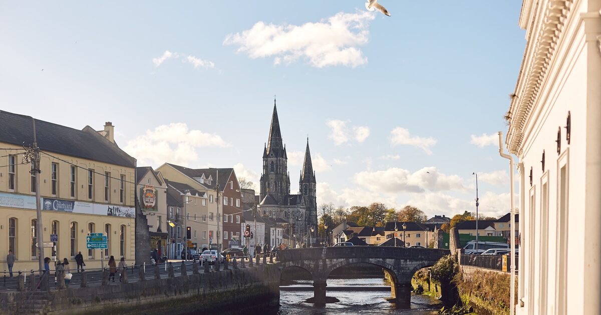 The River Lee in Cork on a sunny day, with old buildings along the quay, a stone bridge and the twin spires of St Fin Barre's Cathedral behind