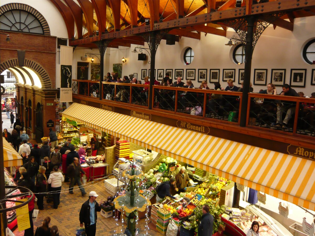 Inside the English Market in Cork: fruit and vegetable stalls under striped awnings, shoppers around the fountain and the café gallery above