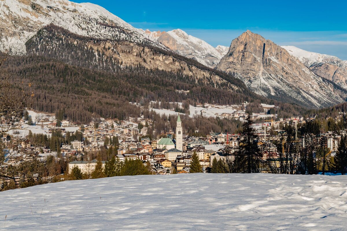 Cortina in winter with the Basilica bell tower among the roofs, snowy meadow in front and the Dolomite peaks behind