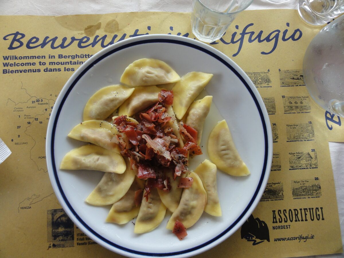 A plate of half-moon casunziei pasta topped with crisp speck, on a paper mat at a mountain hut