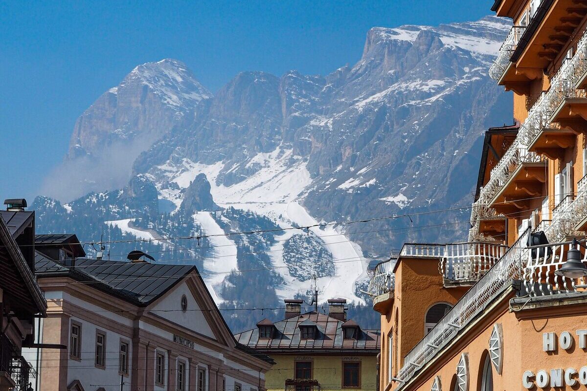 The town hall and a hotel on a sunny street in the center of Cortina, with a snowy mountain and ski runs rising behind the roofs