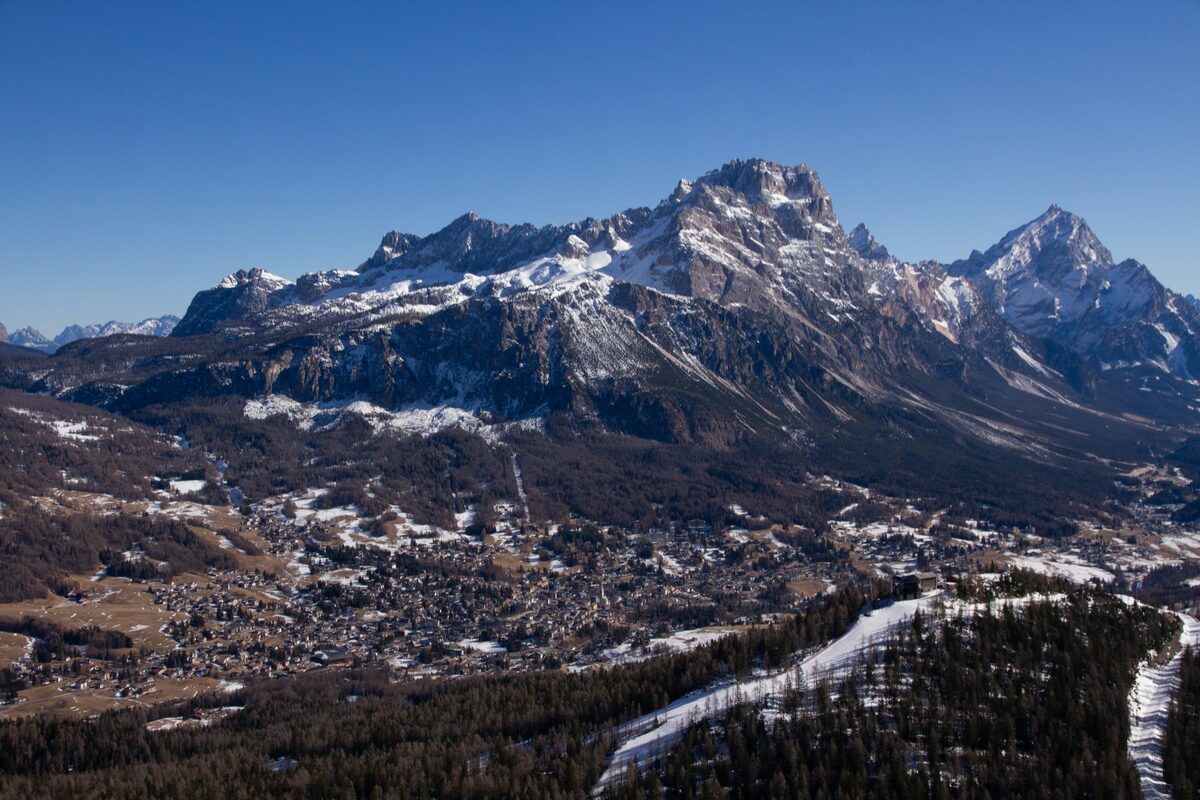 The town of Cortina in its valley below the Dolomite peaks, seen from above, with a snowy ski run cutting through the pine forest in the foreground