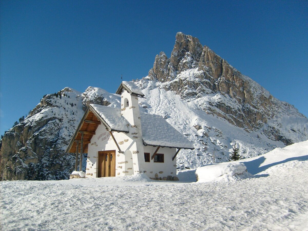 The small white chapel at Passo Falzarego under snow, with the rock peak of Sass de Stria behind it