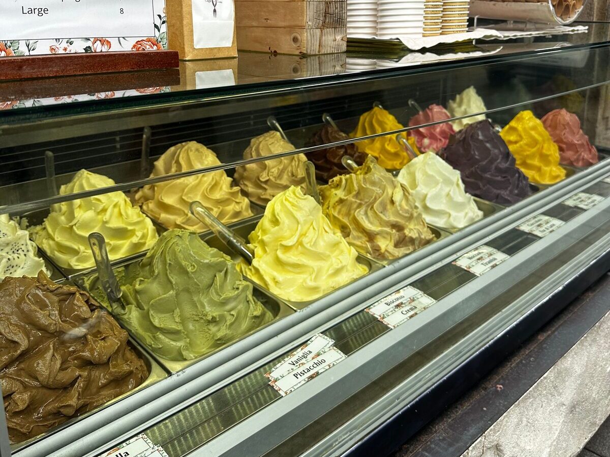 A gelateria counter with rows of pistachio, lemon and chocolate gelato in steel tubs