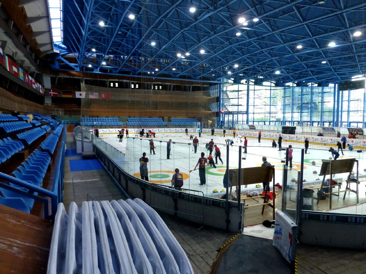 Curlers on the ice of the Olympic Ice Stadium under its blue steel roof, with blue seats and national flags along the side