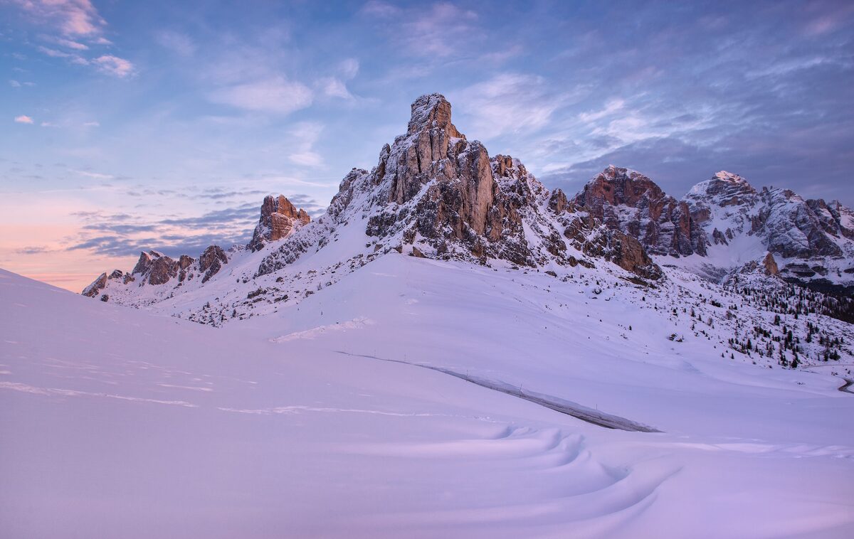 A rocky Dolomite peak above deep snow at Passo Giau, under a pink and blue dusk sky