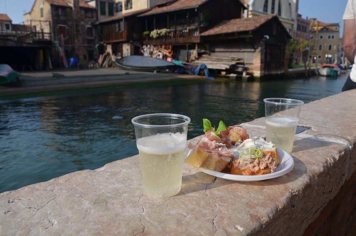 Two glasses of sparkling wine and a plate of cicchetti on a stone ledge beside a Venetian canal