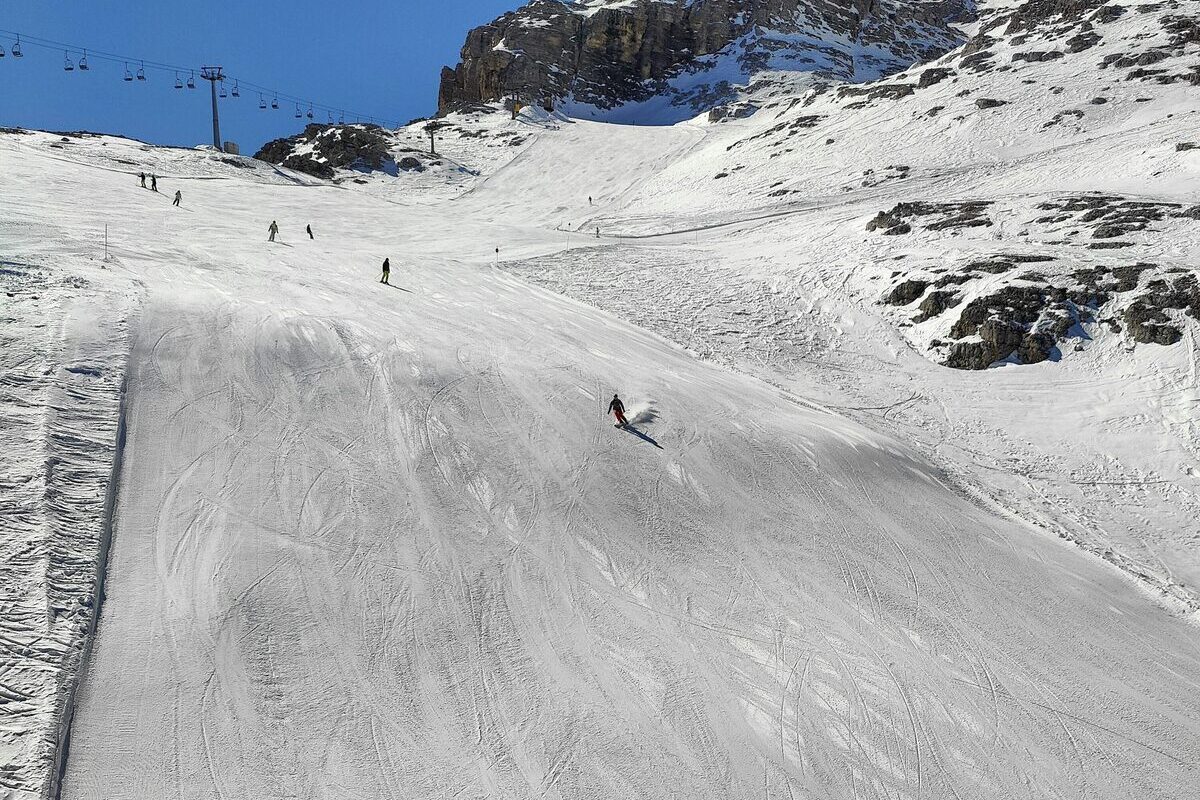 A skier on a wide groomed piste with a chairlift line on the left and rock walls above, under a blue sky