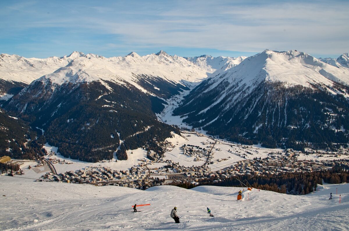 Skiers on a groomed slope above Davos, with the town in the valley and snowy Alps beyond (a Davos ski-area view, not the Kids Slope itself)