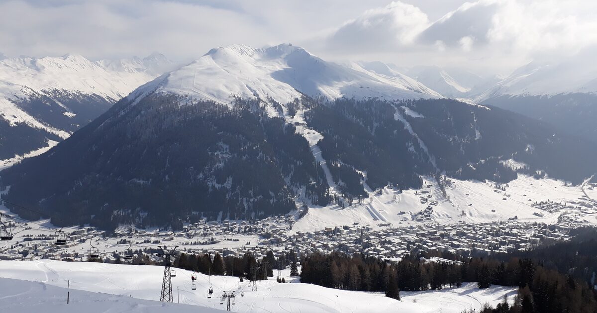 Davos in winter from above: the snowy town in the valley, a steep mountain with ski runs behind it and chairlifts in the foreground