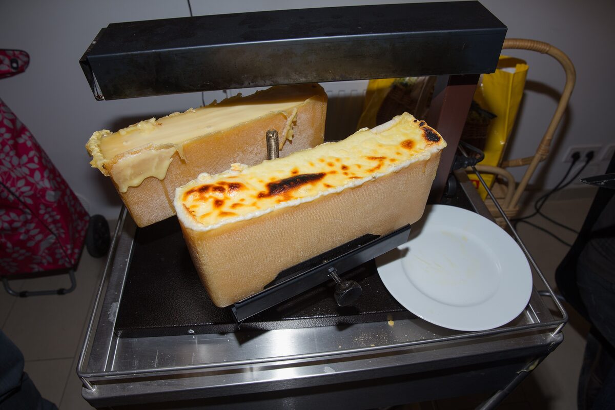 Two half wheels of raclette cheese under an electric grill, the cut faces melted and browned, with a plate ready below