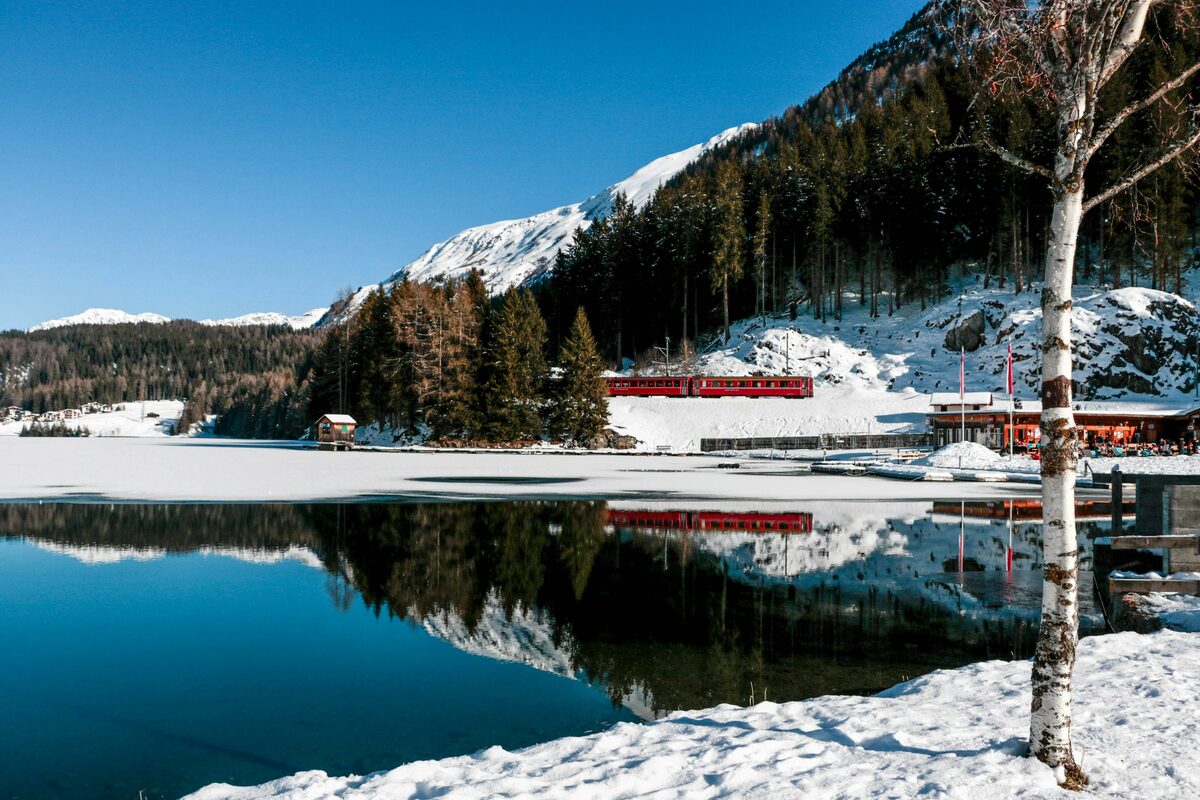 The Davos lake in winter, a red Rhaetian train on the far shore reflected in the water, snowy forest and mountain behind (the lake beside the rink, not the rink)