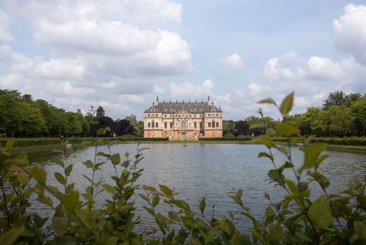 The Palais im Großen Garten across the pond, framed by leaves, under a sky of broken cloud