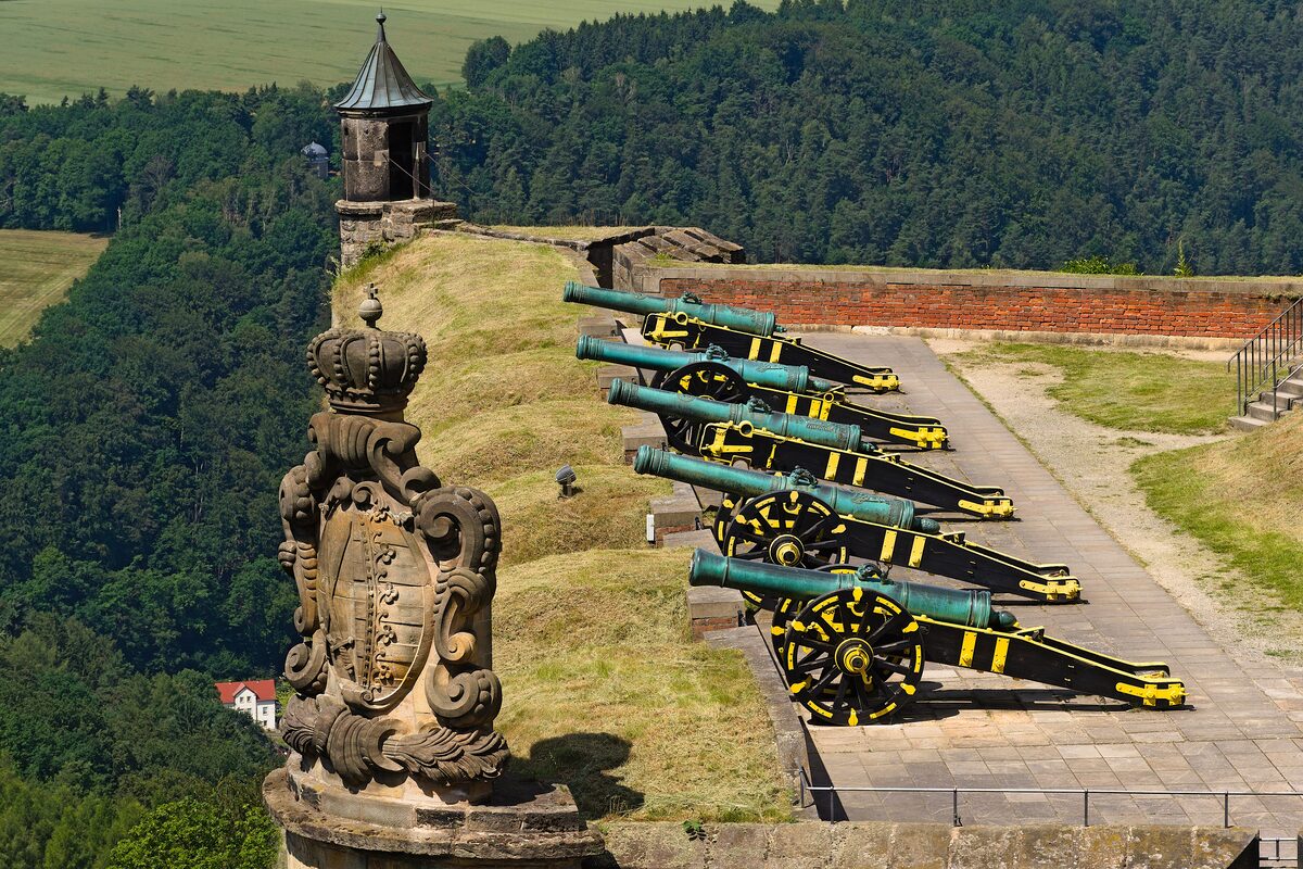 Green-painted cannons on a bastion of Königstein Fortress, with a carved royal coat of arms and forested hills behind