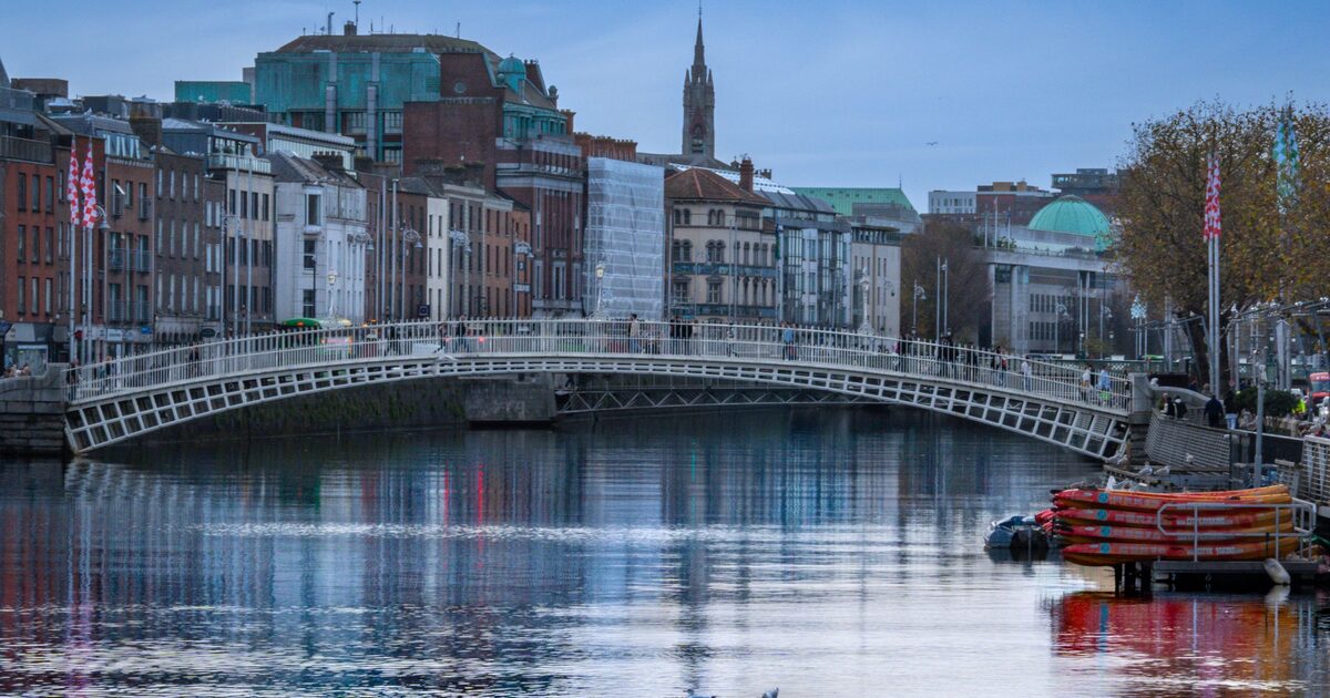 The white iron Ha'penny Bridge over the Liffey at dusk, with old red-brick and stone buildings and a church spire behind it reflected in the river