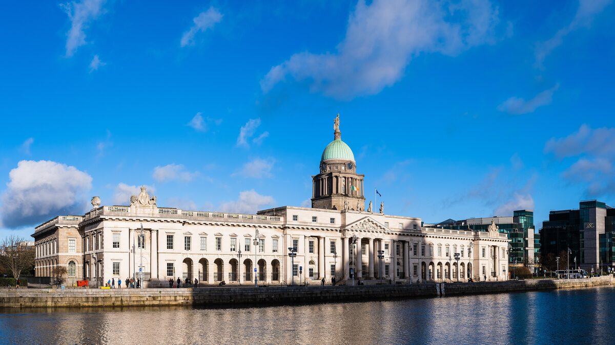 The long white Georgian front of the Custom House with its green dome, reflected in the Liffey under a blue sky