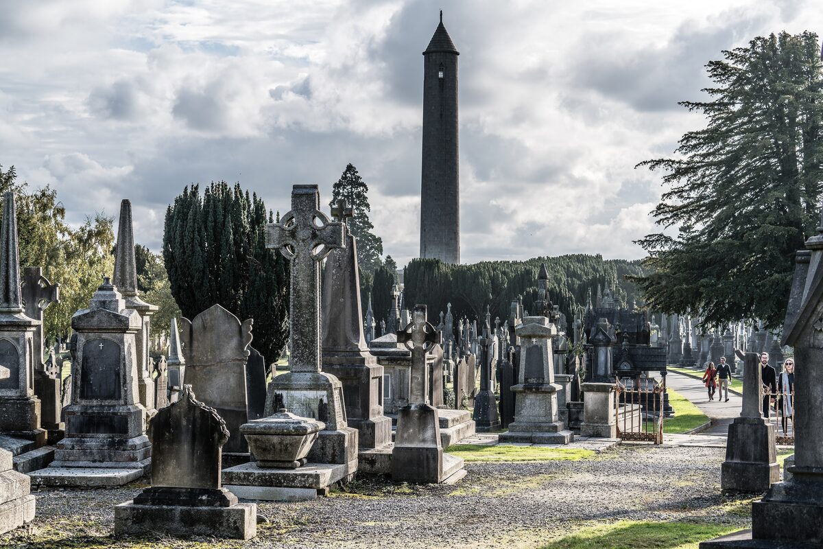 Celtic crosses and Victorian gravestones along a path in Glasnevin Cemetery, with the tall round O'Connell Tower behind