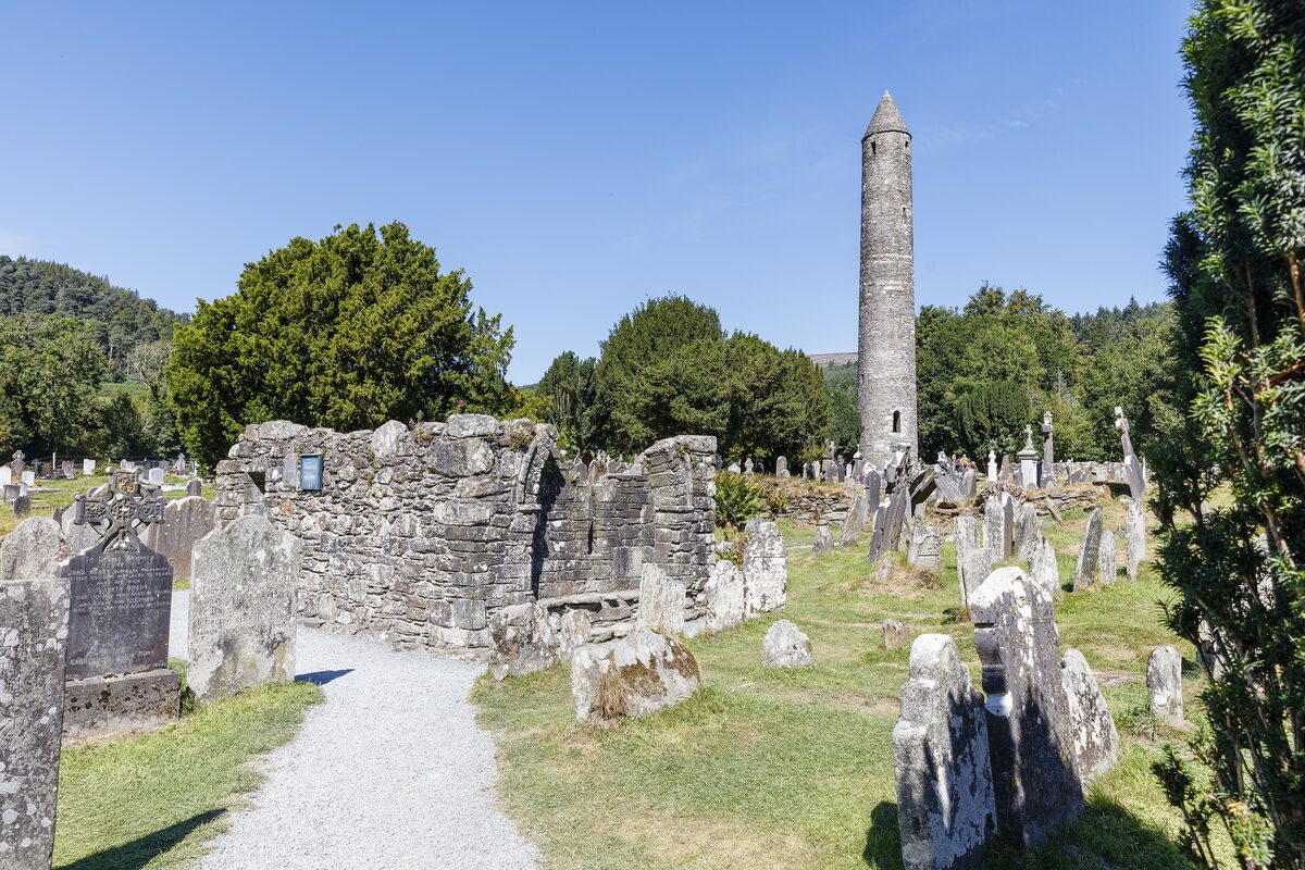 The stone round tower of Glendalough rising above old gravestones, the ruined gable of a church and trees, under a blue sky