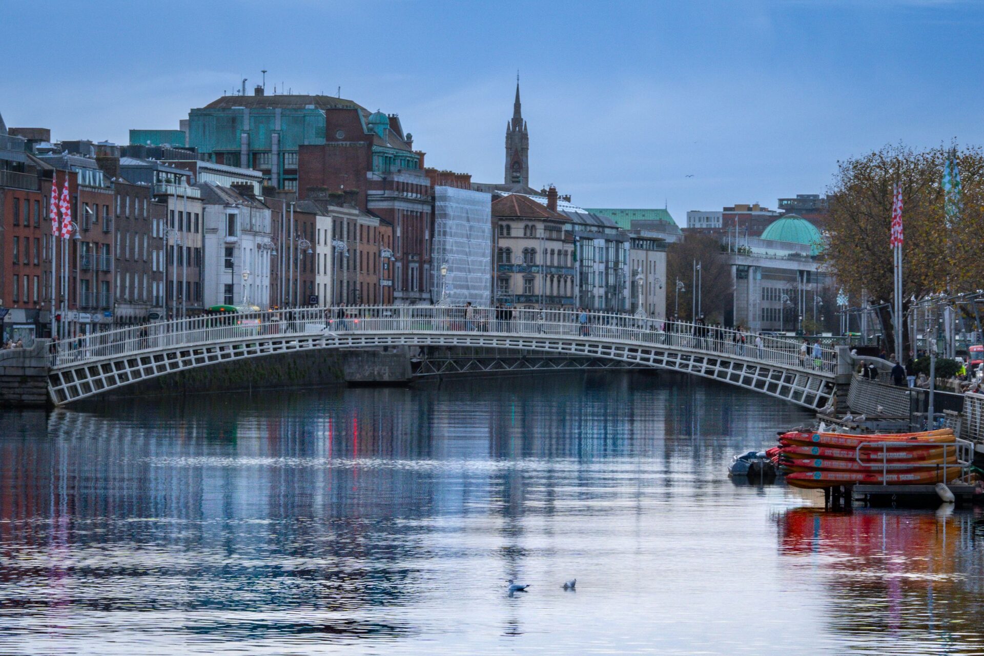 The white iron Ha'penny Bridge over the Liffey at dusk, with old red-brick and stone buildings and a church spire behind it reflected in the river