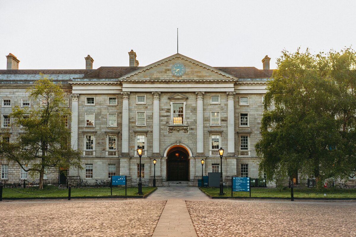 The gray stone front of Regent House at Trinity College Dublin, with lamps along a path across Front Square and trees on either side