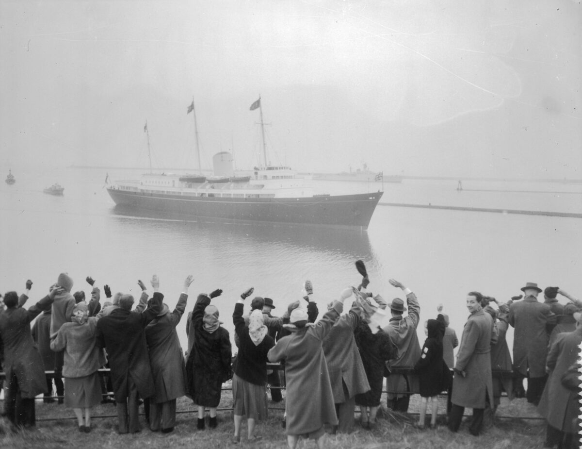 Black-and-white photo of the Royal Yacht Britannia arriving in port in the 1950s, with a crowd waving from the shore