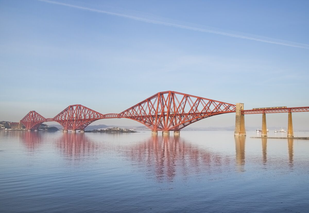 The red steel cantilevers of the Forth Bridge reflected in calm water at dusk