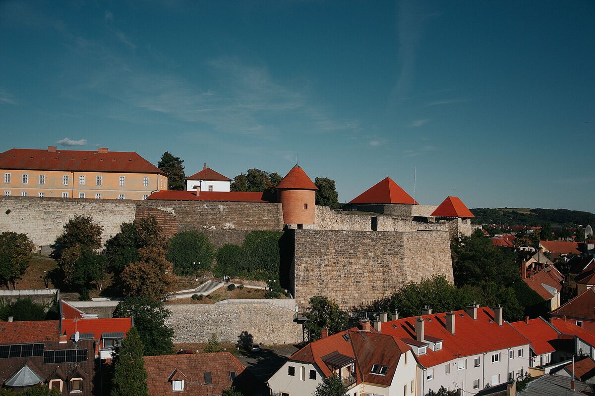 The stone walls and bastions of Eger Castle with red conical tower roofs, rising above the red roofs of the houses below