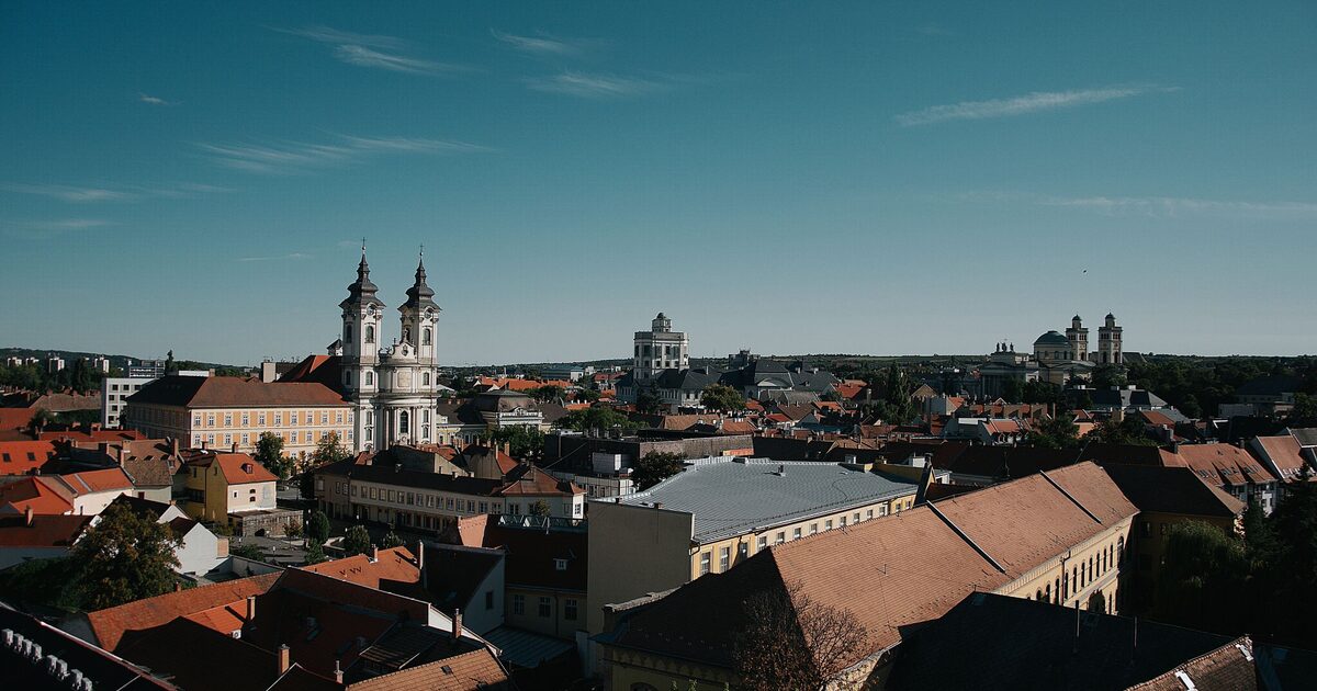 Red-tiled rooftops of Eger under a deep blue sky, with the twin baroque towers of the Minorite Church on the left and the Lyceum and Basilica on the horizon