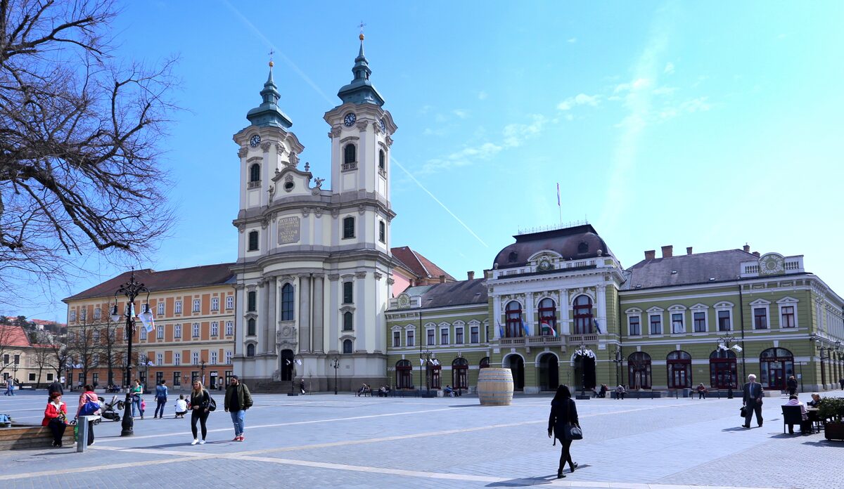 Dobó István Square with the white twin-towered Minorite Church and the green City Hall beside it, people crossing the paved square