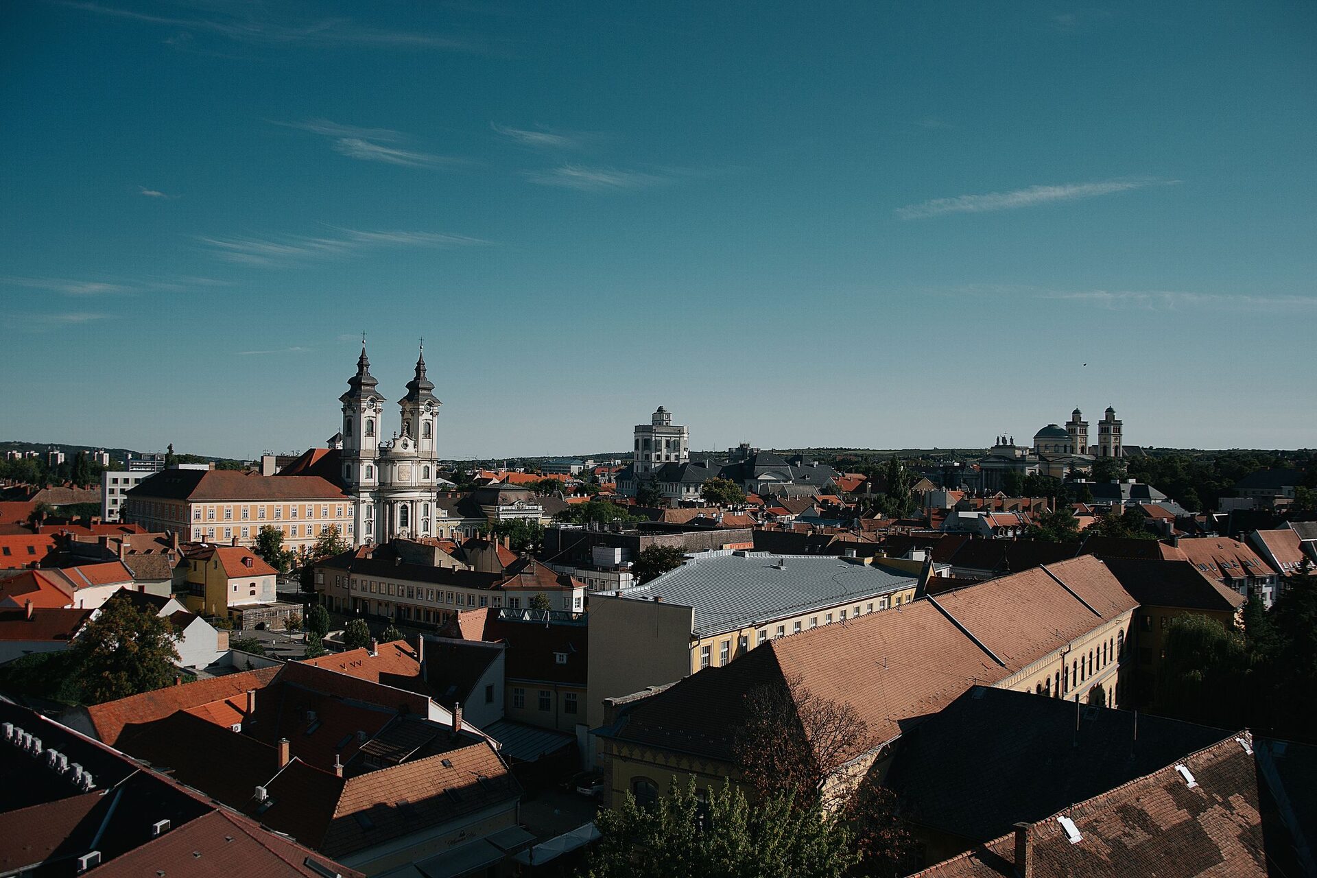 Red-tiled rooftops of Eger under a deep blue sky, with the twin baroque towers of the Minorite Church on the left and the Lyceum and Basilica on the horizon